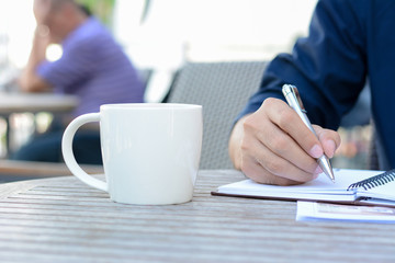 A man hand writing on notebook with coffee cup beside