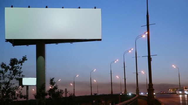 Empty advertising billboard on sidelines of road with traffic at evening