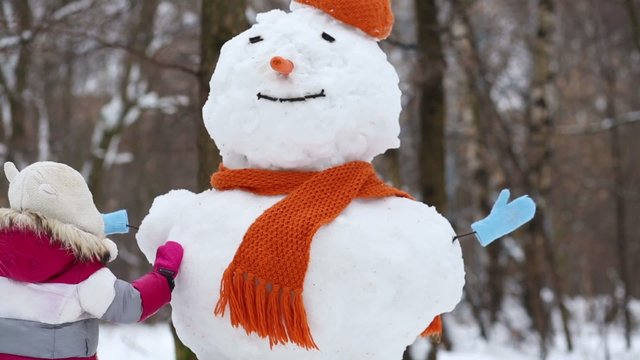 Little girl touch snowman in orange hat and scarf at winter park