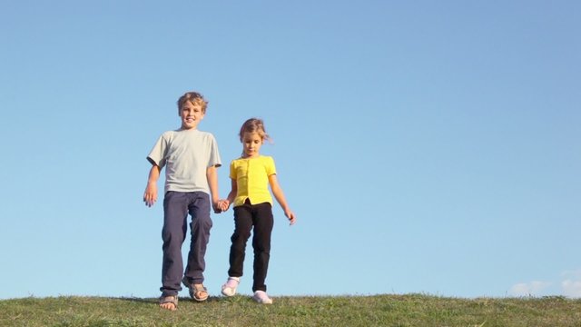 Two kids boy with little girl hold hands and approach from behind of grass hill at sunny summer day