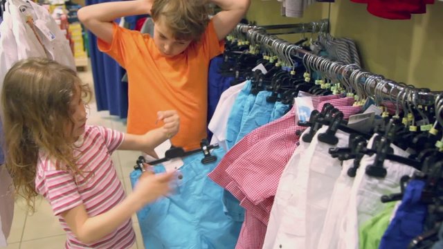 Little girl tries on shorts and her brother watches in clothes shop