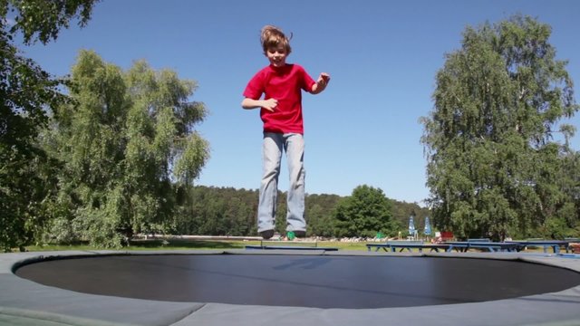 Little boy jumps on trampoline among trees in park at sunny summer day