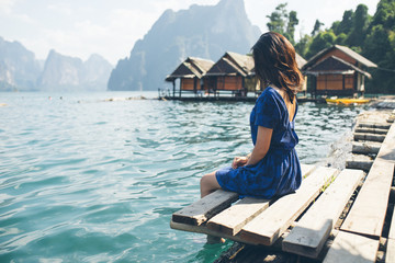 Elegant Asian girl wearing blue dress sitting by huge lake surrounded by limestone cliffs in Khao Sok National Park, Phang Nga province, Thailand