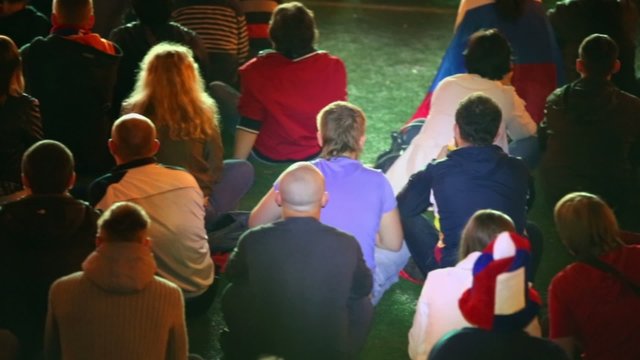 Lot of football fans sit on artificial grass and watch game