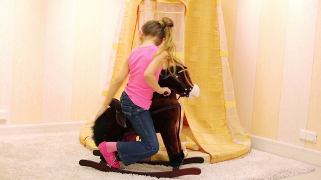 Little girl swinging on a brown rocking horse in light playroom