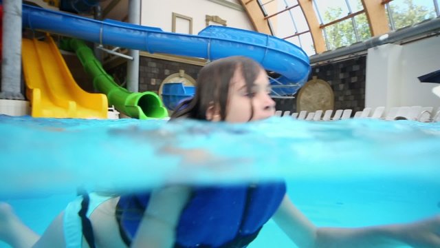 Little girl in lifejacket swims at pool with aqua slides