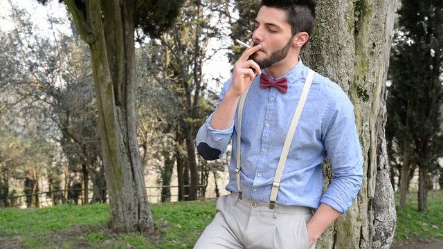 Hipster young man relaxing smoking a cigarette outdoors in a par