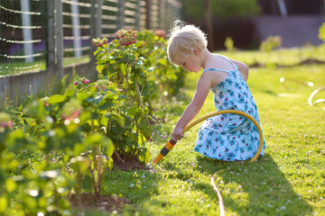 Little girl watering flowers in the garden using flexible hose