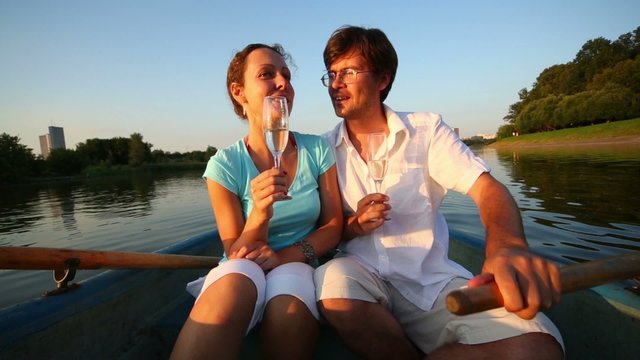 woman and man sits in boat with glasses of wine and talk on pond