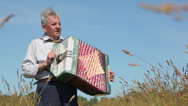 Old man in shirt plays on accordion in field