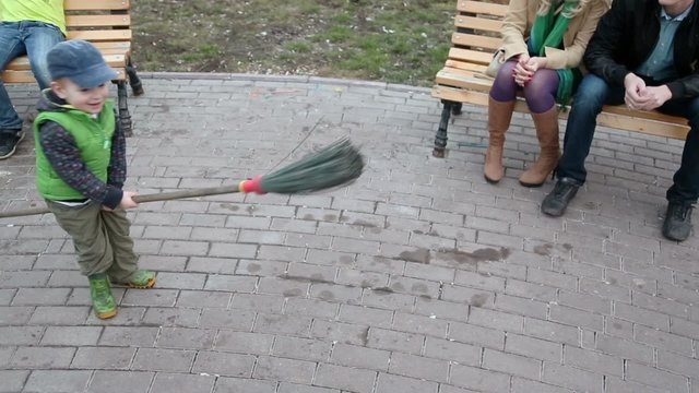 Little boy plays with broom near his parents which sit on bench