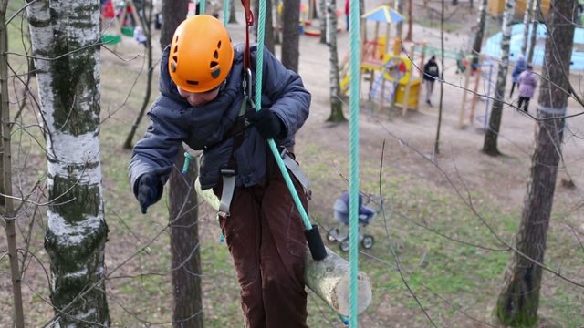 Happy boy in helmet climbs at rope way among trees in park