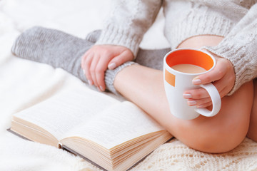 Soft photo of a woman in a cozy sweater on the bed with the old book and a cup of tea with milk in their hands