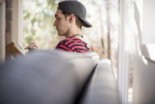 Man wearing a baseball cap backwards, sitting on a sofa, looking at a digital tablet.