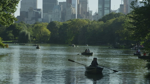 People in rowboats on central park lake with the New York skyline behind them.