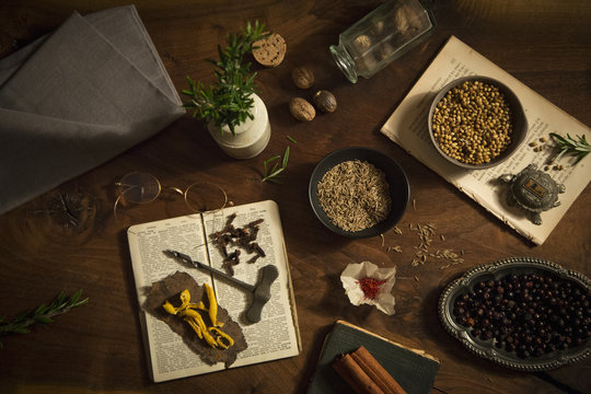 A table spread with a variety of spices and aromatics, coriander, nutmeg, saffron, cloves and rosemary and two old recipe books. 