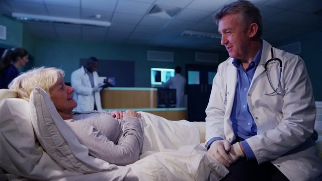 Caring Hospital Doctor Chatting At The Bedside Of Elderly Female Patient At Nigh