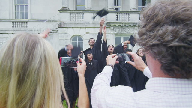Happy students on graduation day stand in a line & pose for photographs outdoors
