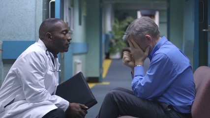 Worried man sitting alone in a hospital waiting area is given bad news by doctor