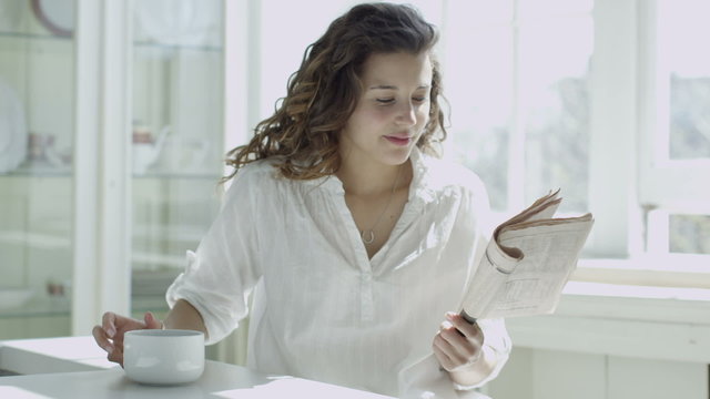 Attractive young single woman relaxing by the window and reading a newspaper