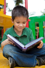Cute primary school aged boy reading book outdoors at playground