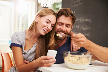 Young Couple Eating Breakfast Whilst Using Mobile Phones