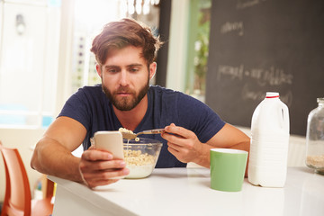Young Man Eating Breakfast Whilst Using Mobile Phone