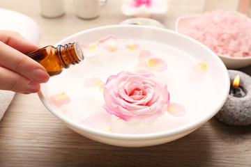 Female hand with bottle of essence and bowl of spa water with flowers on wooden table, closeup