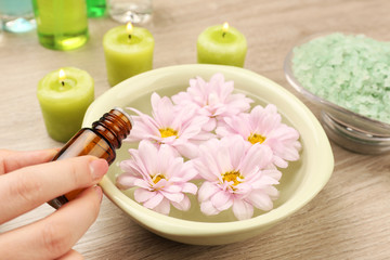 Fototapeta na wymiar Female hand with bottle of essence and bowl of spa water with flowers on wooden table, closeup