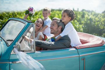 A newlywed couple is driving a convertible car with their kids