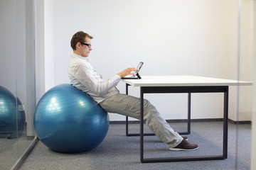 young man on stability ball at desk working  with tablet