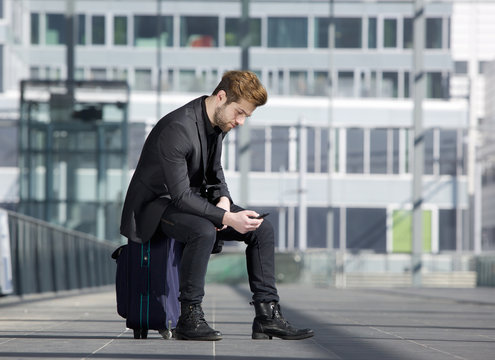 Male traveler sitting on suitcase reading text message