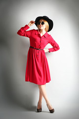Expressive young model in red dress, black hat and sunglasses on gray background