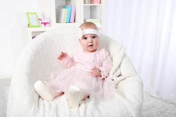 Fototapeta na wymiar Cute baby girl in pink dress sitting in arm-chair, on home interior background