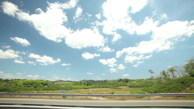 View from car window of road and countryside, with blue sky and clouds in background