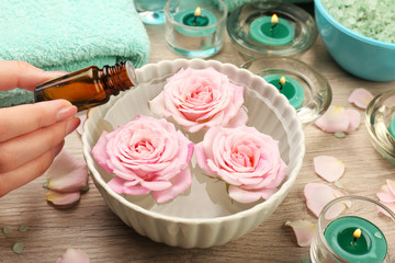Female hand with bottle of essence and bowl of spa water with flowers on wooden table, closeup