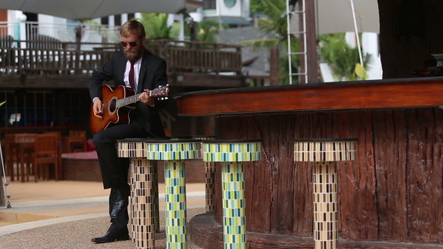 bearded man in suit and glasses plays guitar sitting on bar