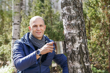 Man in woods collecting birch sap to steel mug