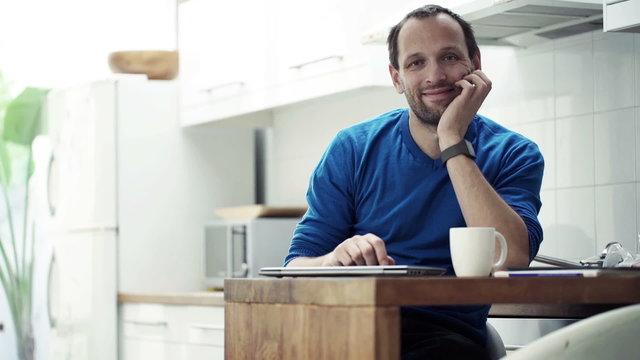 Portrait of happy man with laptop sitting by table in kitchen 