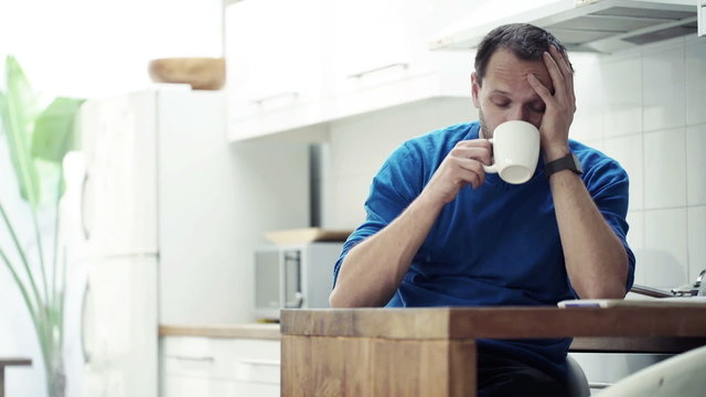 Sad, tired man drinking coffee by table in kitchen at home