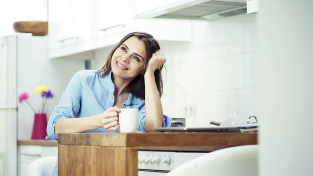 Happy woman drinking coffee sitting by table in kitchen