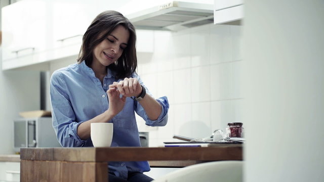 Young, pretty woman with smartwatch sitting by table in kitchen