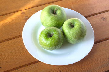 Three ripe green apples on white plate on wooden table