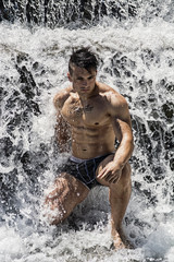 Attractive muscular shirtless young man under small waterfall