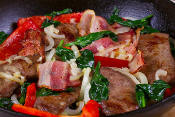 Frying pan with fried meat, bacon, red pepper and spinach