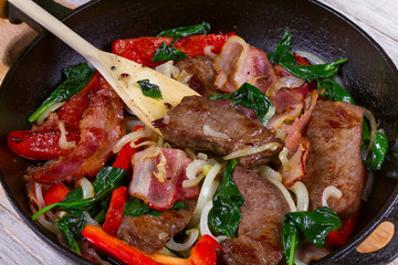Frying pan with fried meat, bacon, red pepper and spinach