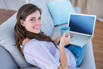 Fototapeta premium Pretty brunette using her laptop with cup of coffee on couch