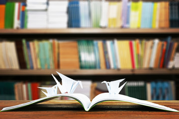 Open book with paper cranes on bookshelves background