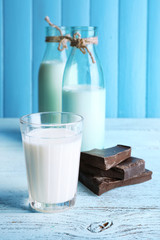 Glass and bottles of milk with chocolate chunks on color wooden planks background
