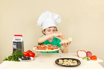Little boy in chefs hat puts a grated cheese on pizza crust